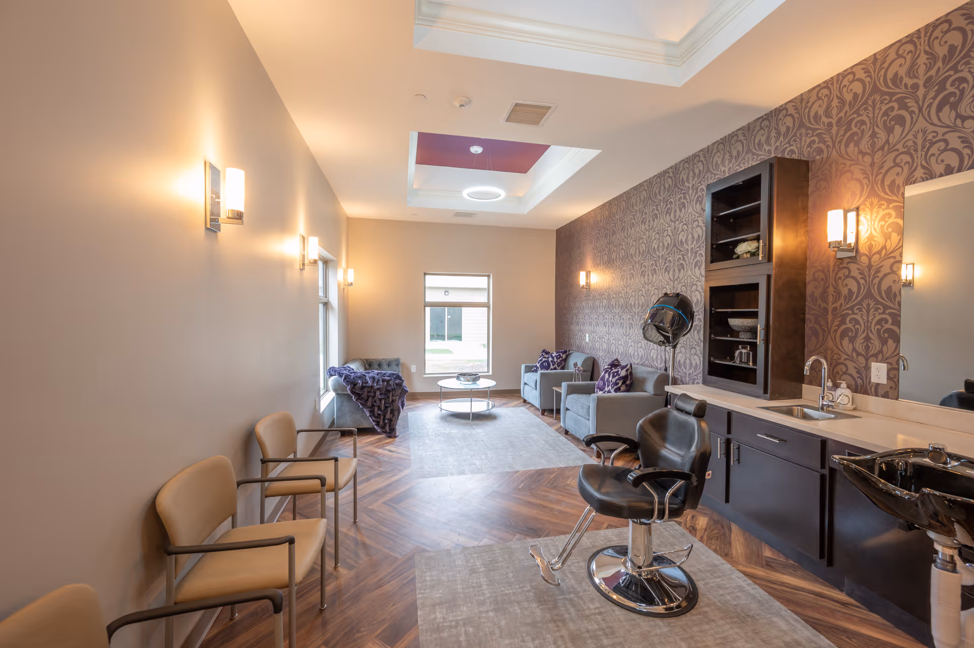 A well-lit interior room with a salon chair and hair dryer station on the right side, a sink, and dark cabinetry. The back wall has patterned wallpaper and two armchairs with purple pillows and a small round table between them. On the left side, there are three beige chairs lined up against a plain wall with wall-mounted lights. The floor has a wood pattern with two area rugs, and a skylight with a circular light fixture is visible on the ceiling.