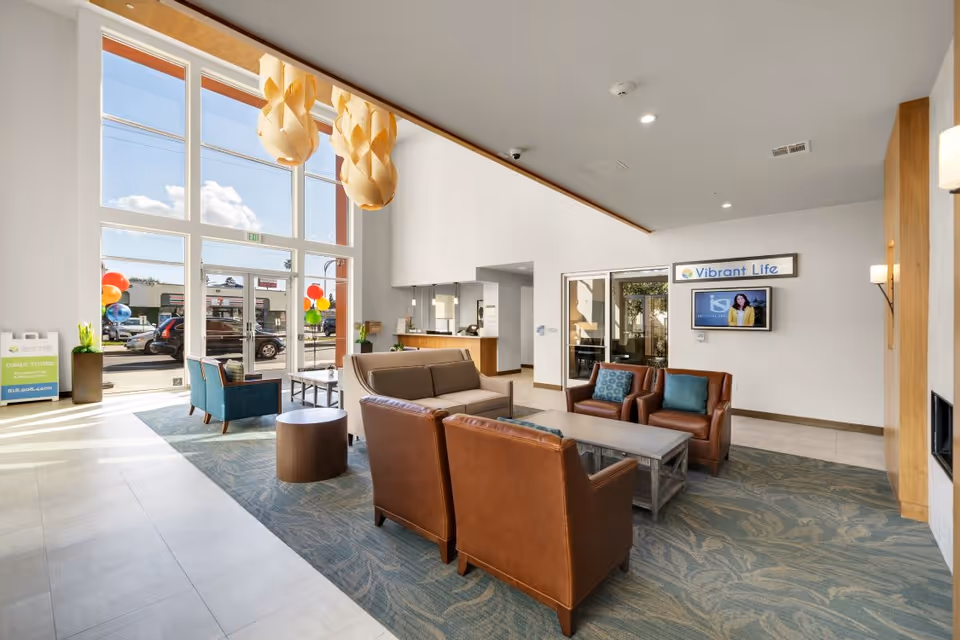 A bright and spacious senior living facility lobby with large floor-to-ceiling windows letting in natural light. The seating area includes a beige sofa, two brown leather armchairs, and a blue armchair arranged around a rectangular coffee table on a patterned rug. Modern pendant lights hang from the ceiling, and a sign on the wall reads 'Vibrant Life' above a mounted TV. Outside, colorful balloons are visible near the entrance.