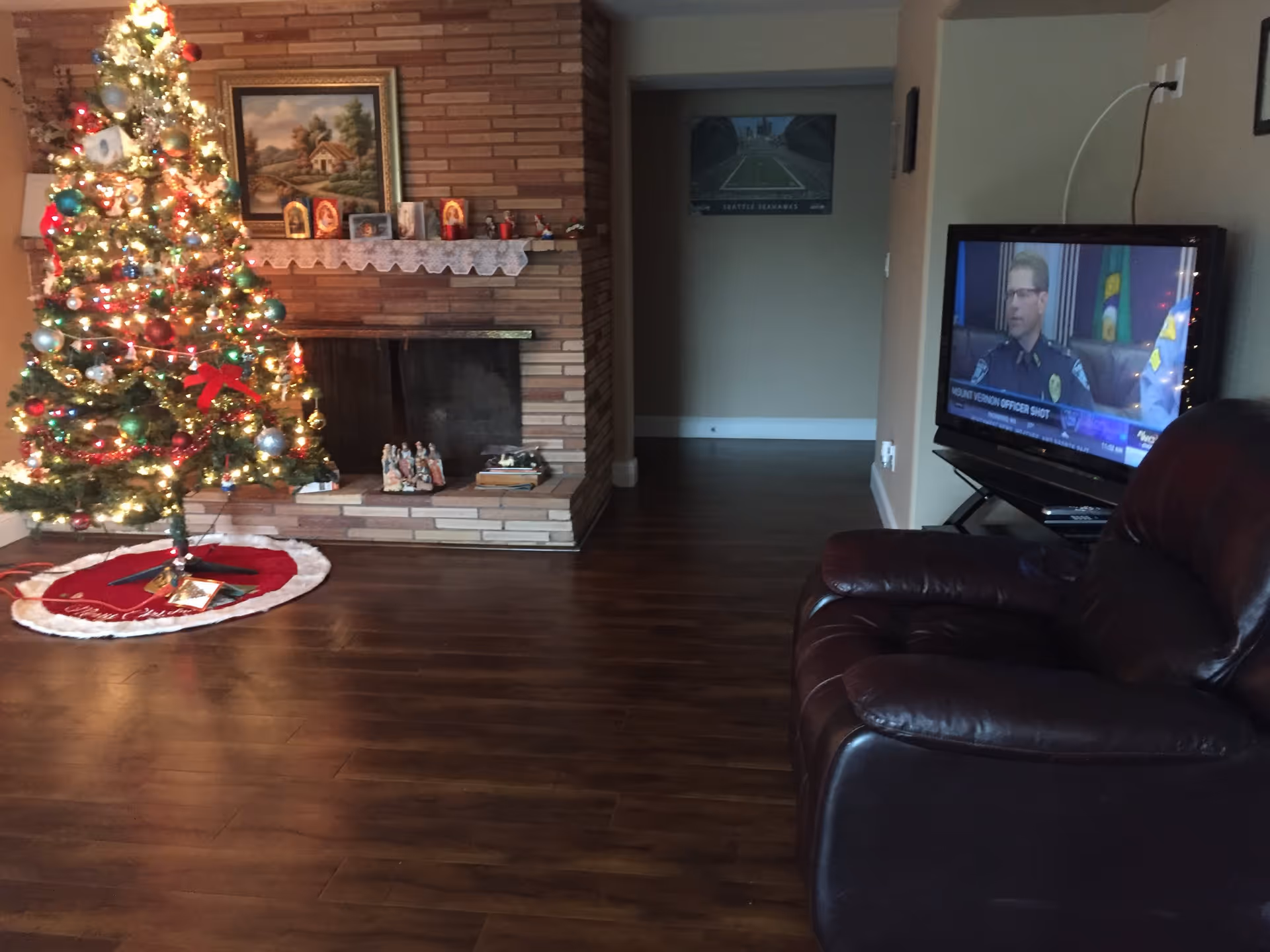 A cozy living room decorated for Christmas with a lit Christmas tree on the left side near a brick fireplace. The fireplace mantel has framed pictures and small decorations. On the right side, there is a dark leather recliner chair facing a television that is turned on, showing a news broadcast with a police officer. The room has wooden flooring and neutral-colored walls.