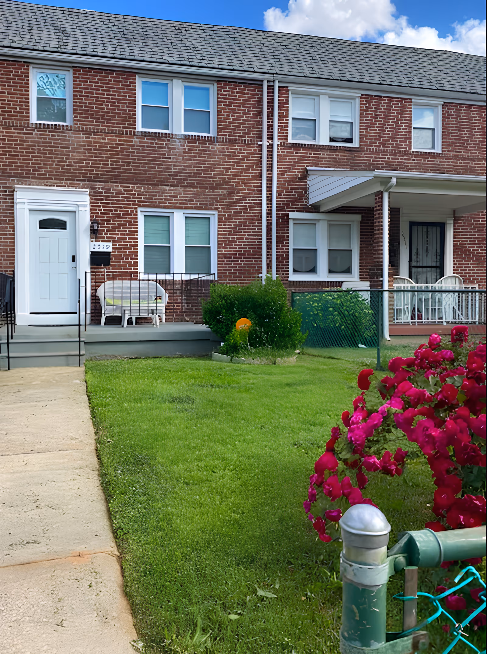 Front view of a red brick townhouse with white framed windows and doors. There is a small porch with a white bench on the left side and a covered porch with white chairs on the right side. The lawn is green and well-maintained with bright pink flowers in the foreground near a green fence post. The sky is partly cloudy.