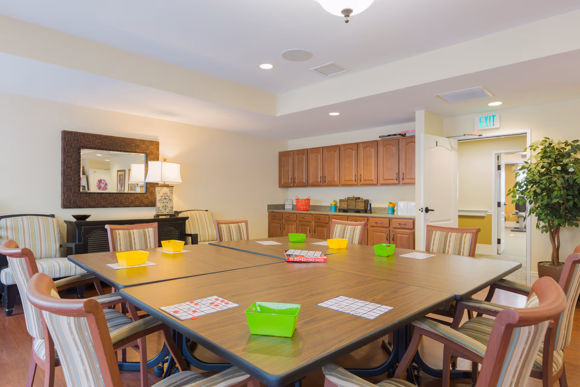 A well-lit activity room with a large square table surrounded by wooden chairs with striped cushions. On the table are bingo cards, colorful plastic baskets, and a box of Pokeno game. The room has wooden cabinets along one wall, a large mirror above a sideboard with a lamp, and an exit door leading to a hallway. A potted plant is near the door.