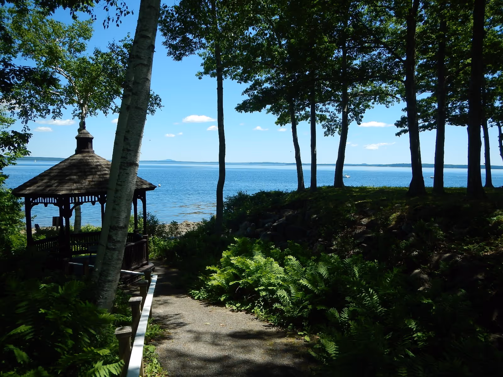 Wooded shoreline path leading to a gazebo overlooking a calm blue lake under a clear sky.