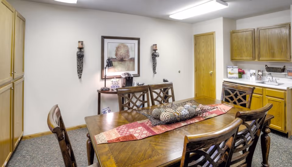 A dining area with a wooden table and six matching chairs. The table has a decorative runner and a centerpiece with patterned spheres. The room features wooden cabinets, a small side table with a coffee maker, a framed picture on the wall, and two wall-mounted candle holders. The flooring is carpeted and the walls are light-colored.