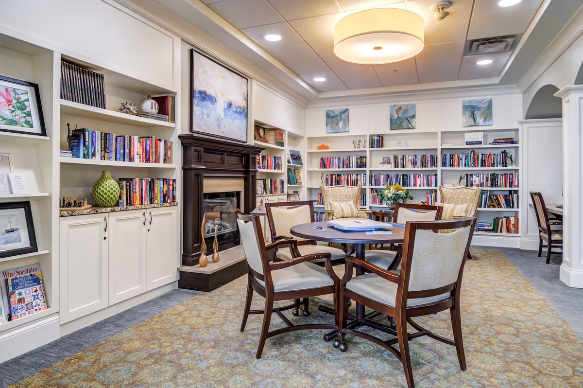 A cozy library or reading room with built-in white bookshelves filled with books and decorative items. There is a dark wood fireplace with a painting above it, a round table with four wooden chairs with cushioned seats in the center, and two armchairs with patterned upholstery near the bookshelves. The room has a patterned carpet and a large circular ceiling light.