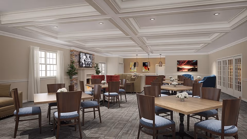 A spacious and well-lit common area in Glenmont Abbey Village featuring multiple wooden tables with chairs, small flower arrangements on each table, a fireplace with a mounted TV above it, comfortable armchairs, and colorful artwork on the walls. The room has a coffered ceiling with recessed lighting and large windows with white curtains.