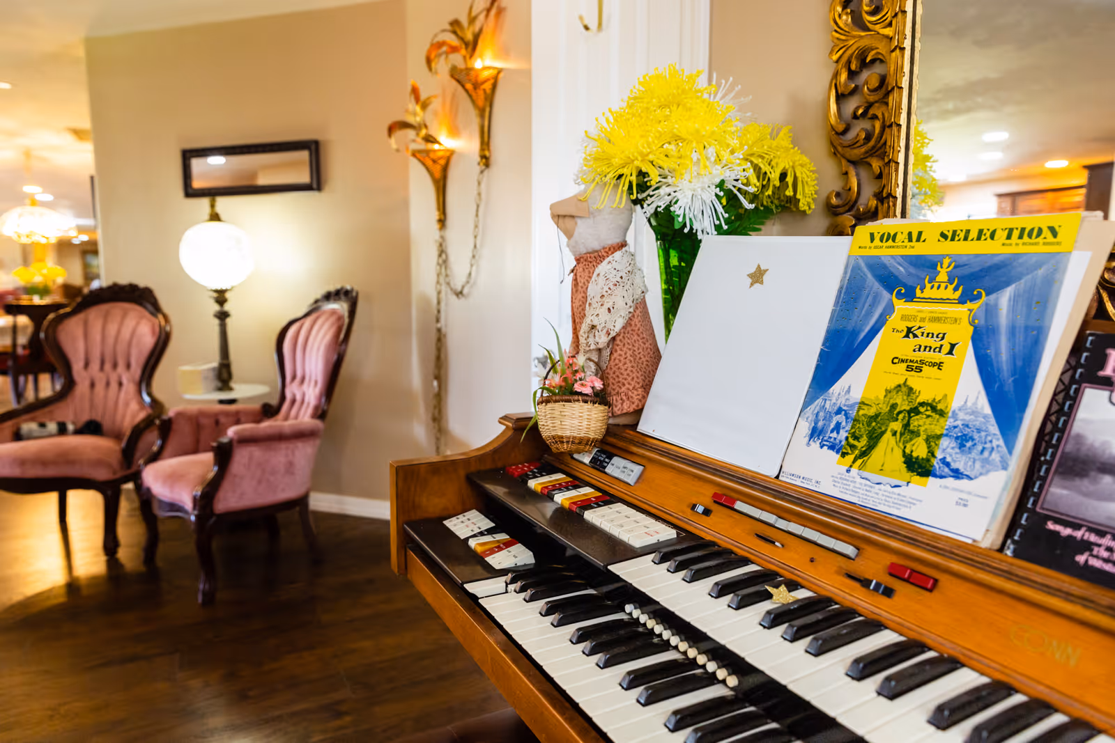 Warm common room interior showing an electric organ with sheet music and flowers in the foreground and upholstered vintage chairs and lamps in the background.