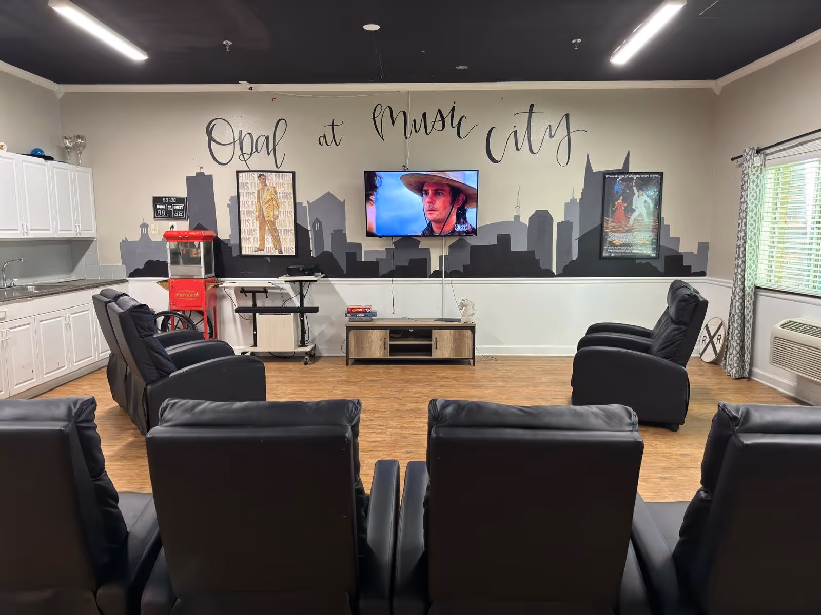 A common area with black leather recliners arranged facing a wall-mounted TV. The wall behind the TV has a mural of a city skyline with the words 'Opal at Music City' written above. There is a popcorn machine, a scoreboard, and framed posters on the wall. White cabinets and a countertop are on the left side, and a window with patterned curtains is on the right.