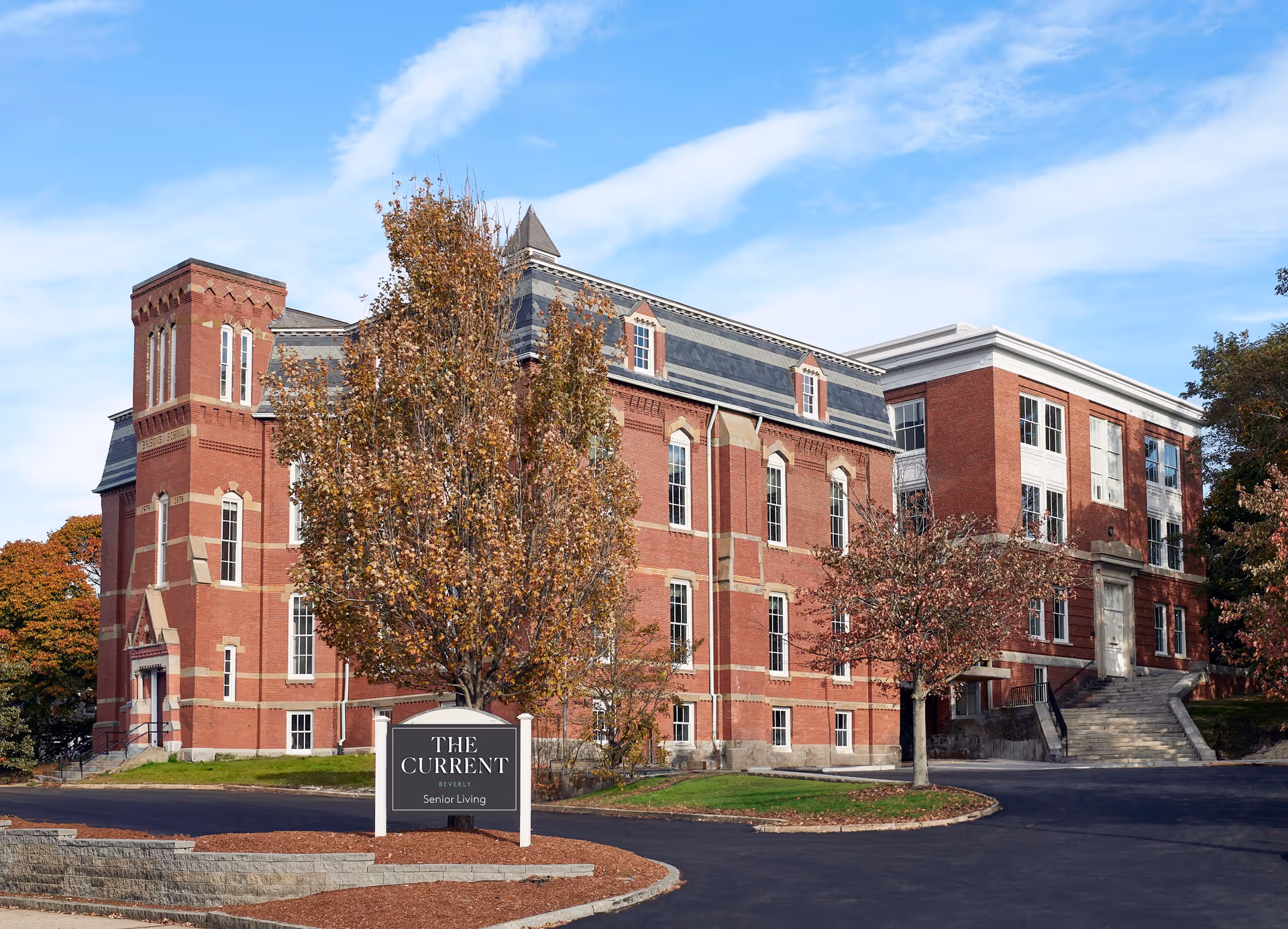Exterior view of a large, historic red brick building with multiple windows and a slate roof under a blue sky with some clouds. In front of the building, there is a sign that reads 'The Current Beverly Senior Living' surrounded by landscaped mulch and trees with autumn foliage.