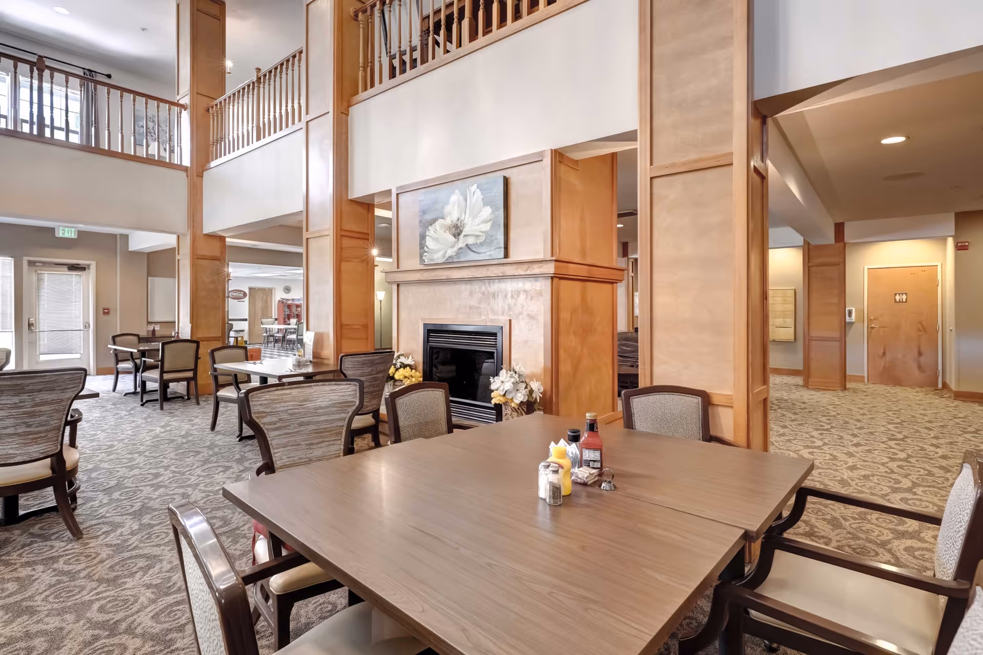 A spacious dining area in an assisted living facility featuring multiple wooden tables and cushioned chairs. The room has a high ceiling with a second-floor balcony railing visible. There is a fireplace with a floral painting above it, and condiment bottles on the tables. The carpet has a patterned design, and there are doors and hallways leading to other parts of the facility.