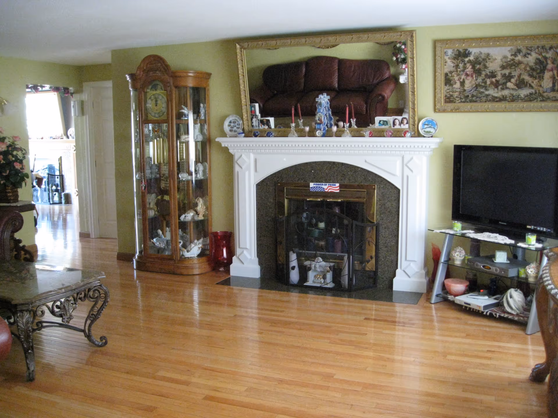 A living room with polished wooden floors, a white fireplace with a decorative screen and various items on the mantel, a large mirror above the fireplace reflecting a brown leather couch, a glass display cabinet with collectibles, a flat-screen TV on a stand with shelves holding electronic devices and decorative items, and a framed tapestry on the wall.