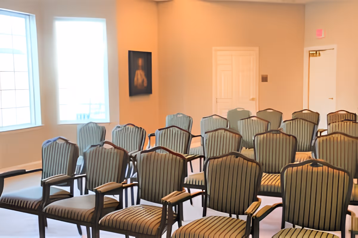 A room with multiple rows of striped upholstered chairs arranged facing forward, large windows letting in natural light, a painting on the wall, and a closed door in the background.