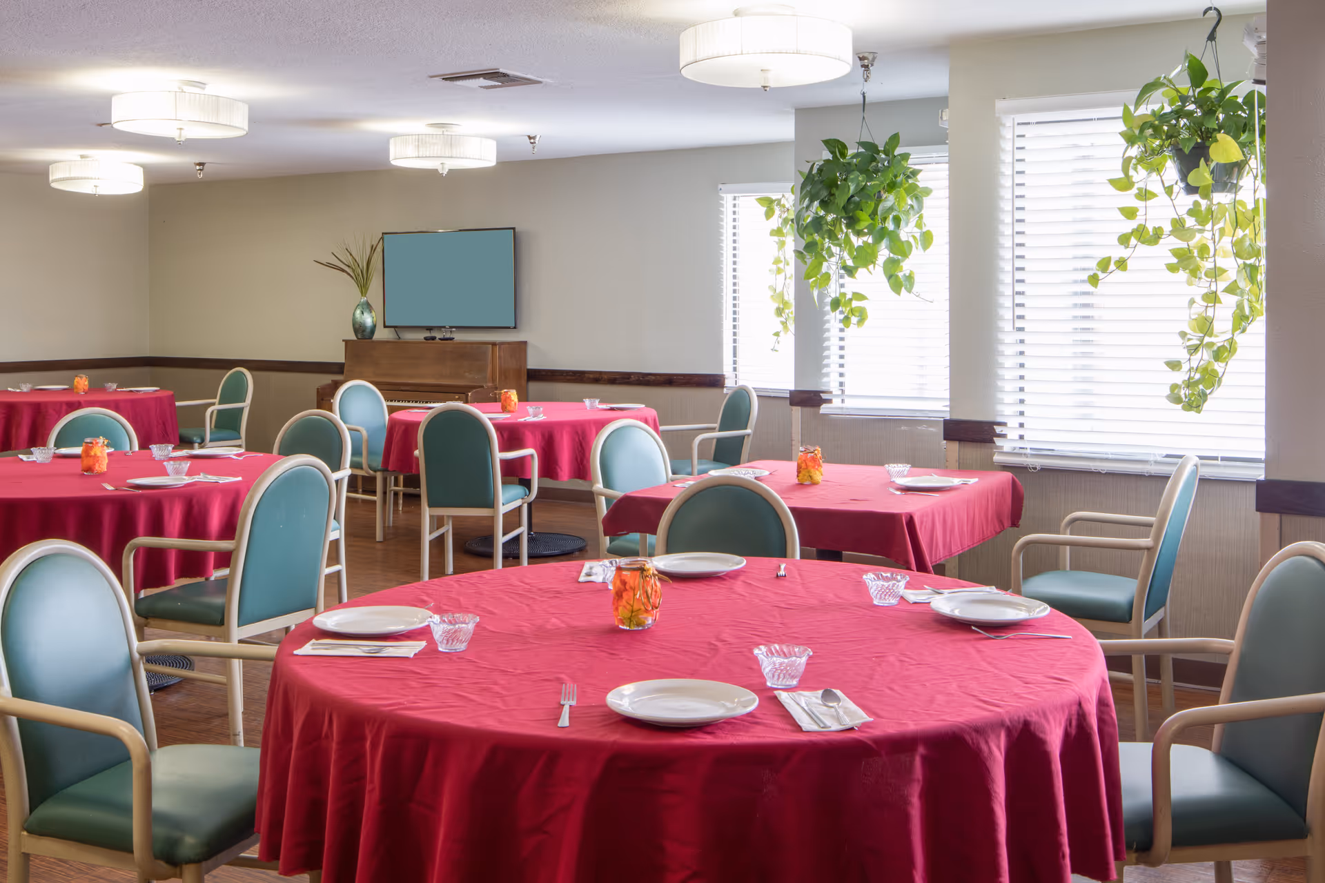 Dining room with round tables covered in red tablecloths, each set with white plates, utensils, and glass bowls. Green cushioned chairs surround the tables. The room has large windows with blinds and hanging green plants. Ceiling lights provide illumination, and a piano with a vase is visible in the background.