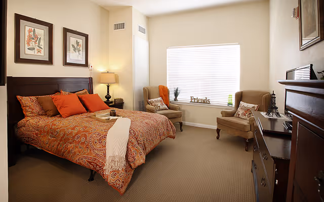 Bright bedroom with a patterned orange bedspread on a wooden bed, two armchairs by a large window, dresser and bedside lamp.