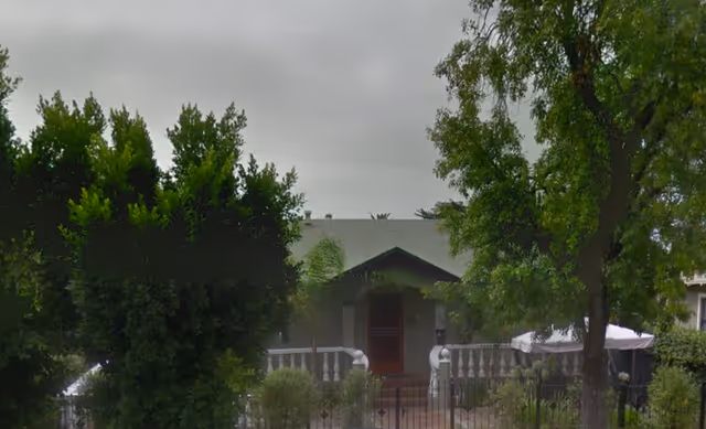 Front of a single-story house with a covered porch, fenced yard, and trees partially obscuring the view.