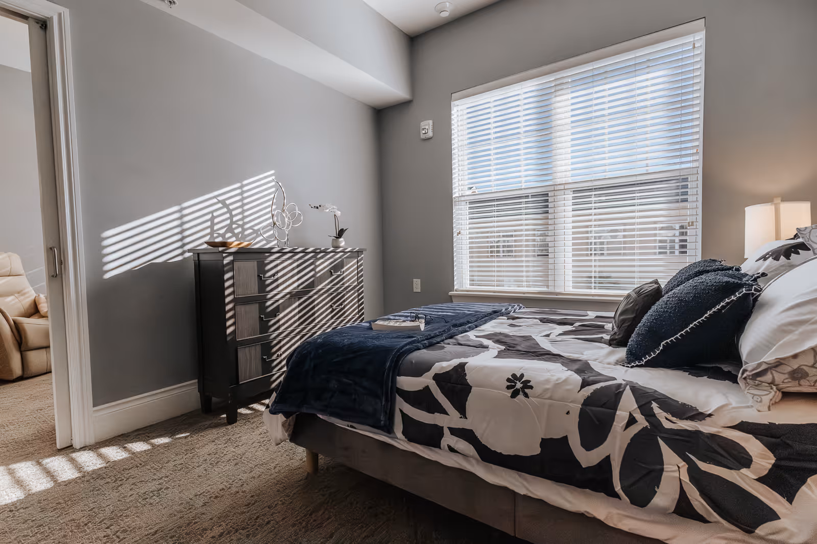 A bedroom with a bed covered in a black and white floral patterned comforter and several pillows. A dark wooden dresser with decorative items on top is against the wall. Sunlight streams through a window with white blinds, casting shadows on the wall and floor. An open doorway reveals a beige recliner chair in an adjacent room.