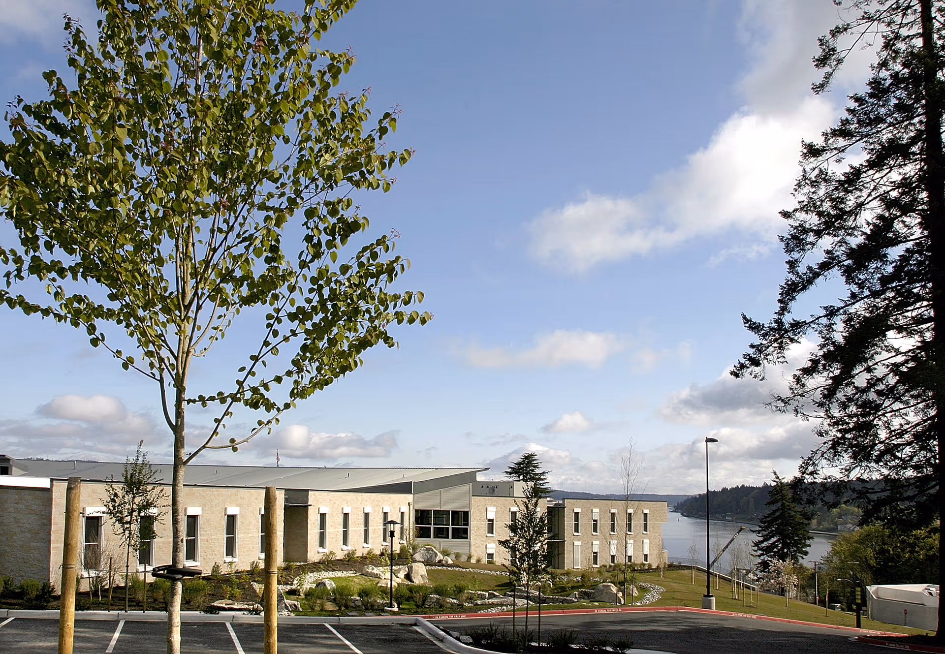 Exterior view of a low-rise beige senior living facility overlooking a waterfront with trees, parking spaces, and a clear blue sky.
