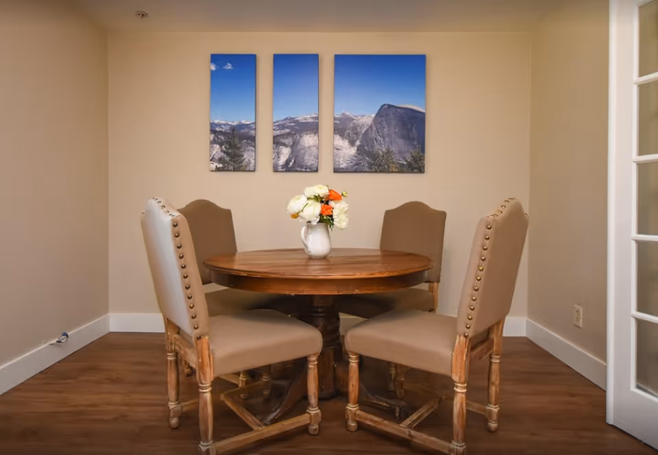 Small dining area with a round wooden table, four upholstered chairs, a vase of flowers, and a three-panel mountain landscape on the wall.