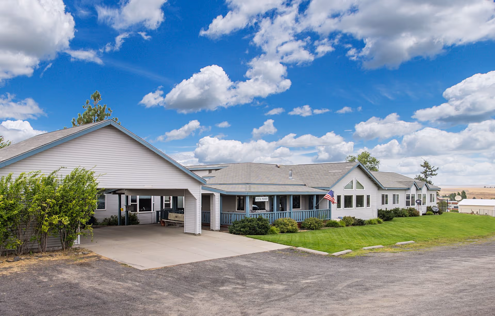 Exterior view of Vista Manor, a single-story building with white siding and a covered entrance. The building is surrounded by green grass and shrubs, with an American flag displayed near the entrance. The sky is blue with scattered clouds.
