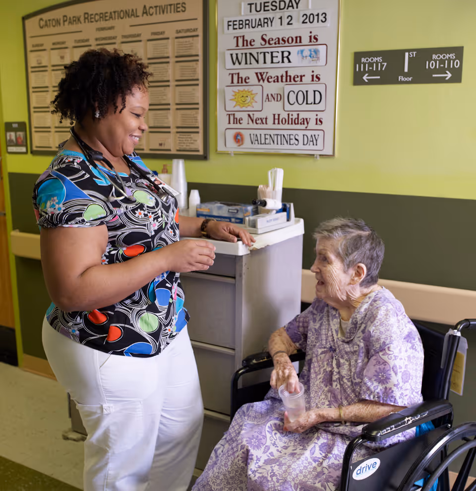 A healthcare worker in colorful scrubs smiles and interacts with an elderly woman seated in a wheelchair holding a plastic cup. Behind them is a bulletin board displaying Caton Park Recreational Activities and a sign indicating the date as Tuesday, February 12, 2013, with information about the season, weather, and upcoming holiday. Directional signs for room numbers are also visible on the wall.