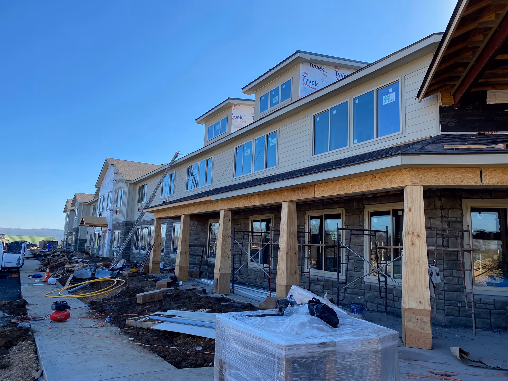 A senior living facility under construction with scaffolding, construction materials, and equipment scattered around the site. The building has multiple windows and a partially completed exterior with stone and siding. The sky is clear and blue.