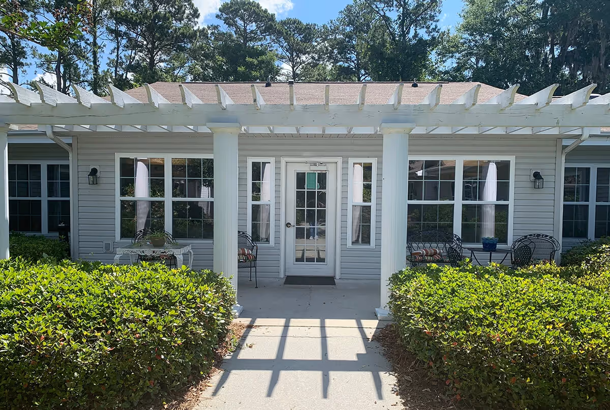 Exterior view of a building at Helena Square with a white pergola supported by two columns over a concrete walkway. The building has multiple windows with white frames and a glass door in the center. There are green bushes on either side of the walkway and outdoor seating with chairs and small tables near the windows. Trees and a blue sky with clouds are visible in the background.