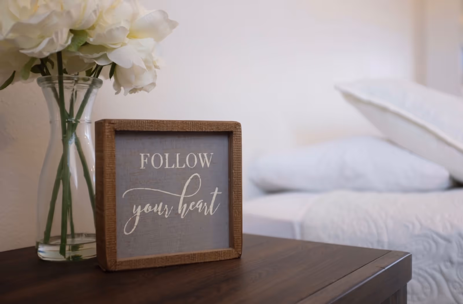 A close-up of a wooden nightstand with a glass vase holding white flowers and a framed sign that reads 'FOLLOW your heart'. In the background, there is a bed with white bedding and pillows.