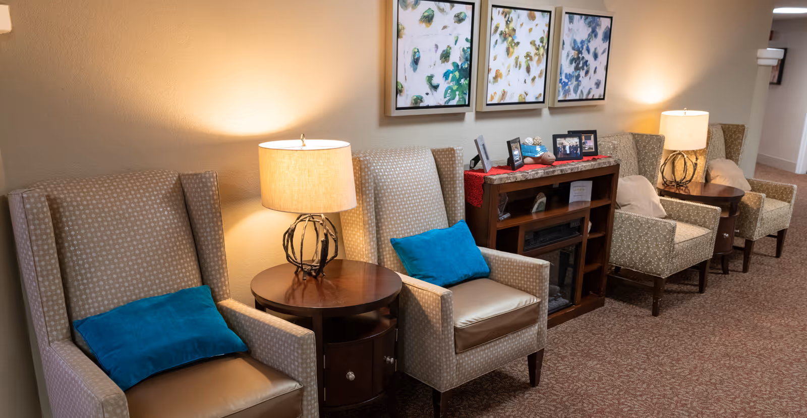 A row of upholstered armchairs with teal accent pillows, side tables and lamps along a carpeted interior hallway with framed artwork on the wall.