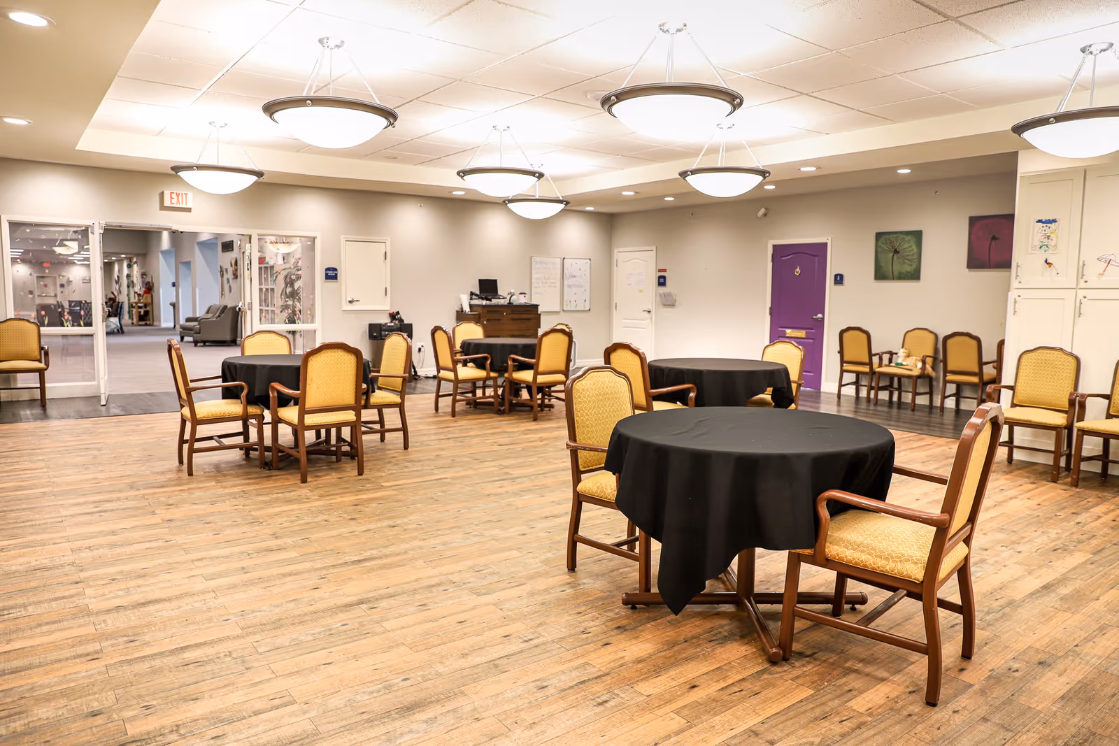 Spacious senior living common/dining room with round tables draped in black cloth and wooden chairs on a wood floor under ceiling lights.