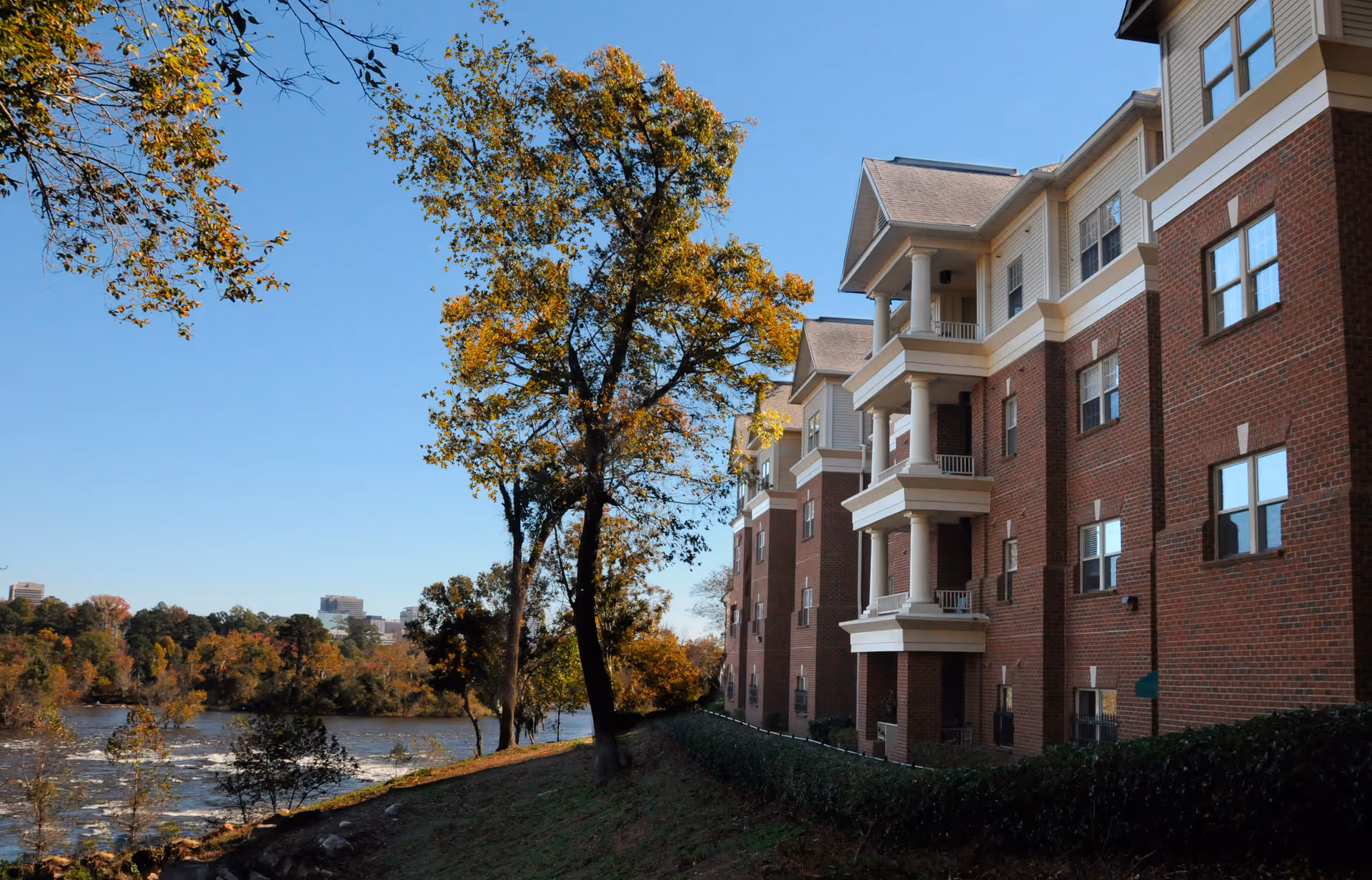 Brick multi-story retirement community building with balconies beside a river and trees under a clear blue sky.