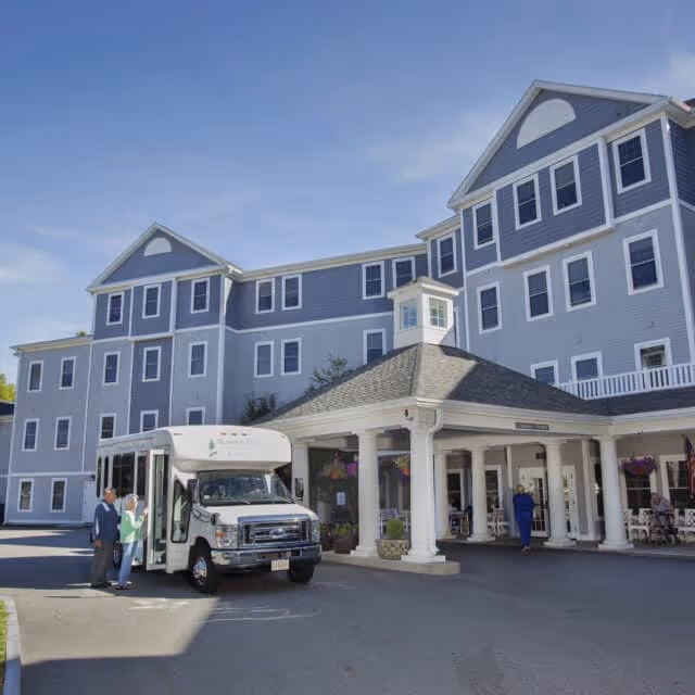 Exterior view of a large, multi-story senior living facility building with gray siding and white trim. A covered entrance with white columns is visible, along with a small shuttle bus parked in front. Several people are standing near the bus and entrance under a clear blue sky.