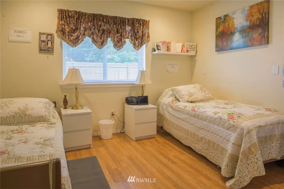 A cozy bedroom with two single beds, each with floral bedspreads. Between the beds are two white nightstands with table lamps and a small trash bin on the floor. A window with floral valance curtains is centered on the back wall. There are framed photos and signs on the walls, including one that reads 'AGNES BED' and another that reads 'JACKIE'S BED'. A colorful painting hangs on the right wall above one of the beds.