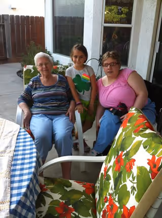 An elderly woman, a young girl, and a middle-aged woman sitting together on a covered patio with floral cushioned chairs and a table with a blue and white checkered tablecloth.