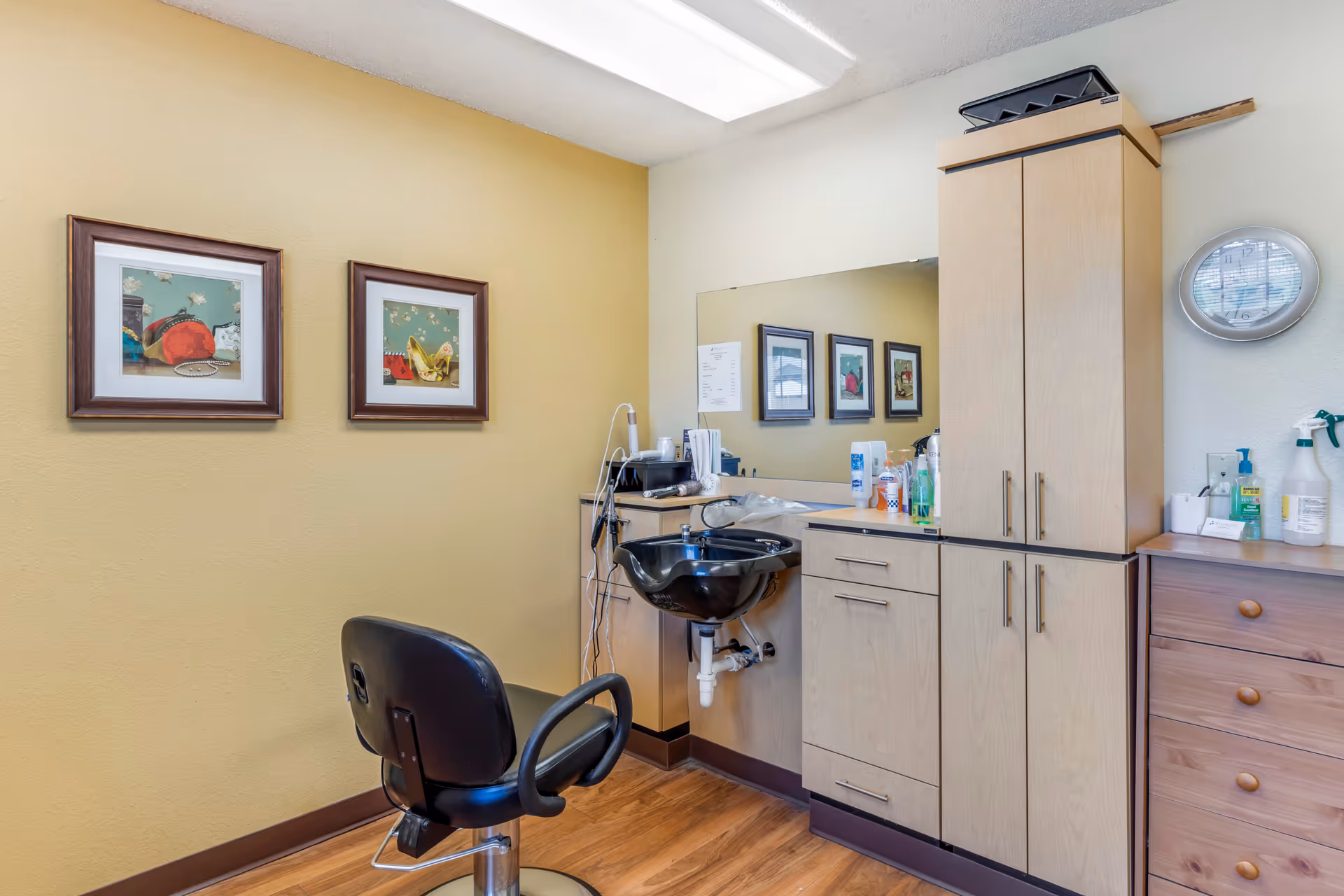 Small salon-style room with a styling chair, wash basin, cabinets, mirror, and framed artwork on a yellow wall.