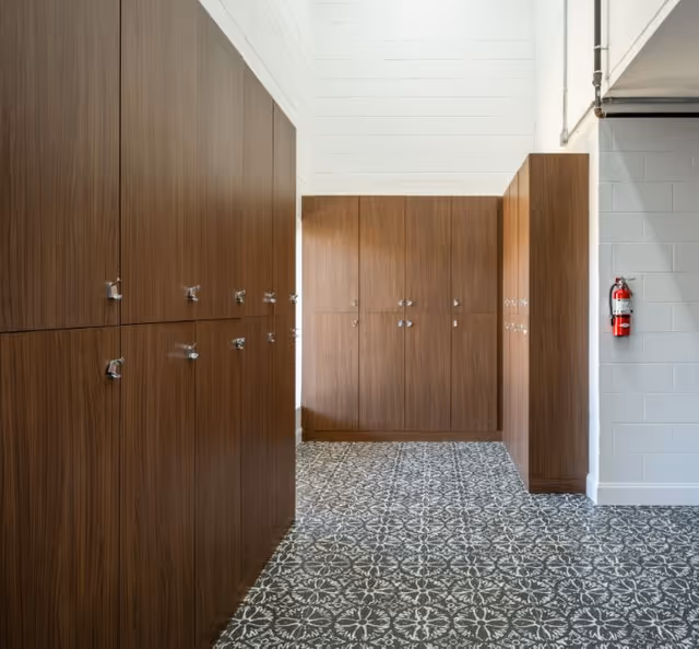 A hallway with multiple wooden lockers on both sides and patterned tile flooring. A fire extinguisher is mounted on the white brick wall to the right.