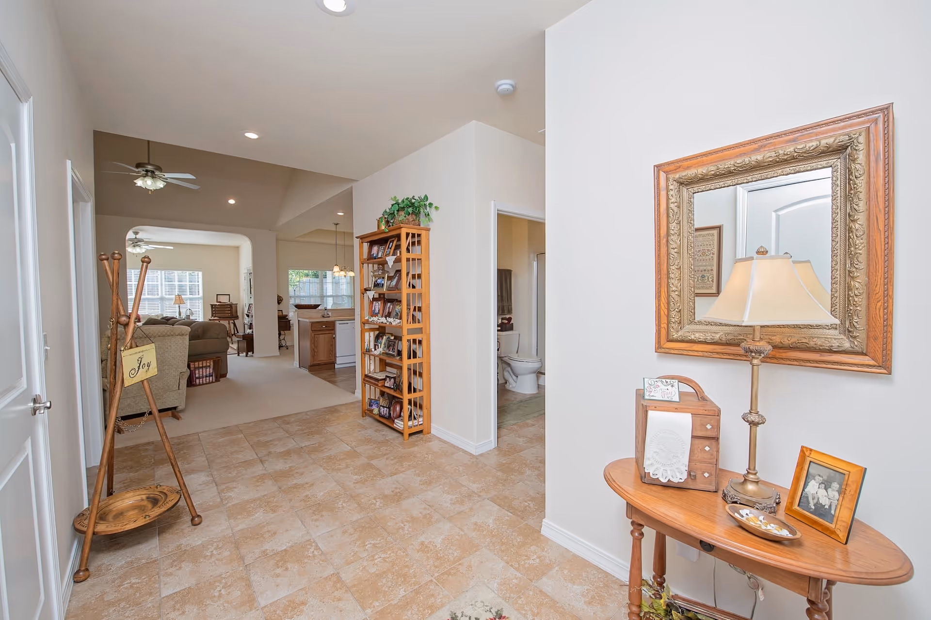Interior view of a senior living facility showing a tiled hallway with a wooden coat rack holding a sign that says 'Joy' on the left. On the right, there is a wooden table with a lamp, framed photo, and decorative items beneath a large ornate mirror. In the background, there is a living room with sofas and ceiling fans, a kitchen area with wooden cabinets and a dishwasher, and a bathroom with a visible toilet.