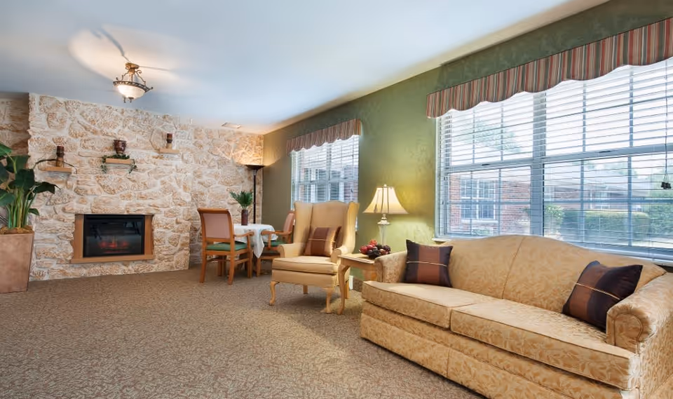 A cozy living room area in a senior living facility with a beige patterned sofa and matching armchair, both adorned with dark brown pillows. A side table with a lamp and decorative fruit bowl sits between the sofa and armchair. Large windows with striped valances allow natural light to fill the room. A stone wall with a built-in electric fireplace and small shelves with plants and candles is visible on the left side. A small dining table with two chairs is positioned near the fireplace, and a tall floor lamp stands in the corner.