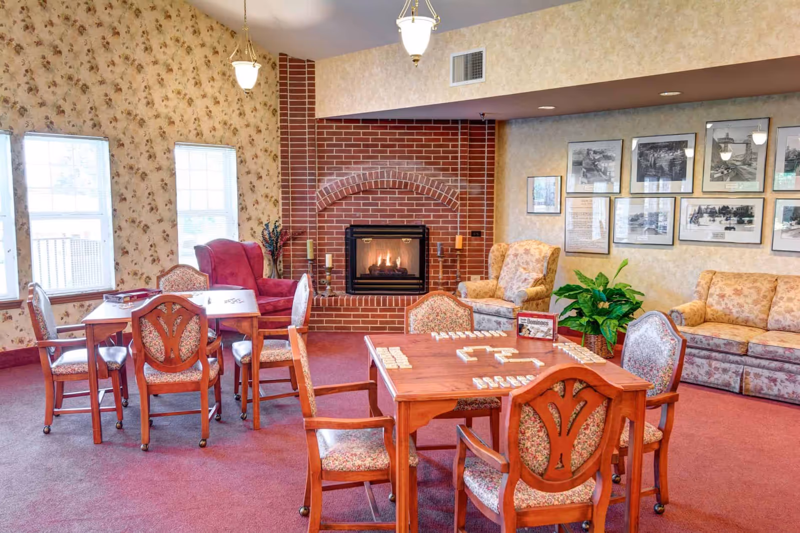 A cozy common room with floral wallpaper and red carpet featuring a brick fireplace with a fire burning. There are two wooden tables with floral upholstered chairs around them, one table set up for a game of dominoes. The room also has a red armchair, two floral armchairs, a floral sofa, framed black and white photos on the wall, and a green potted plant.