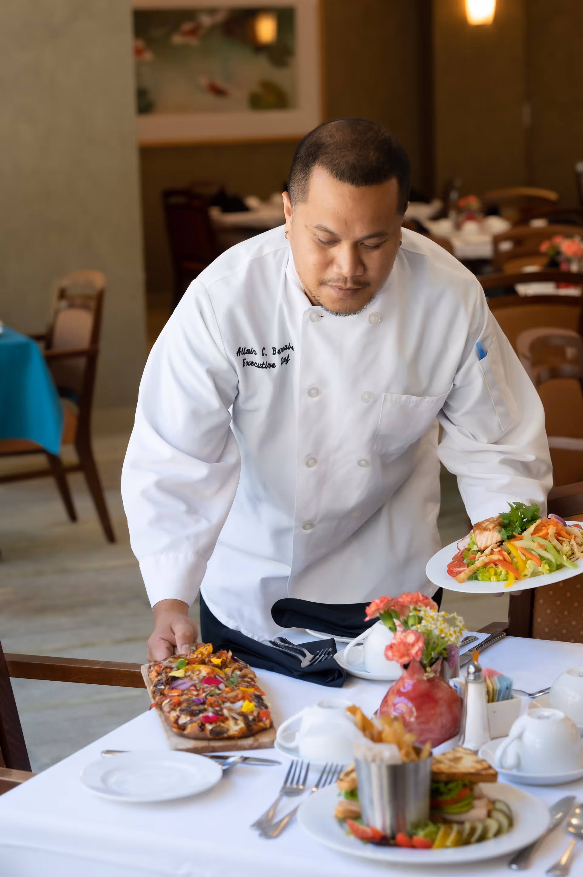 A chef in a white uniform placing a wooden board with a colorful flatbread pizza on a table set with plates, cutlery, and a vase of flowers in a dining room. The chef is also holding a plate with a fresh salad.
