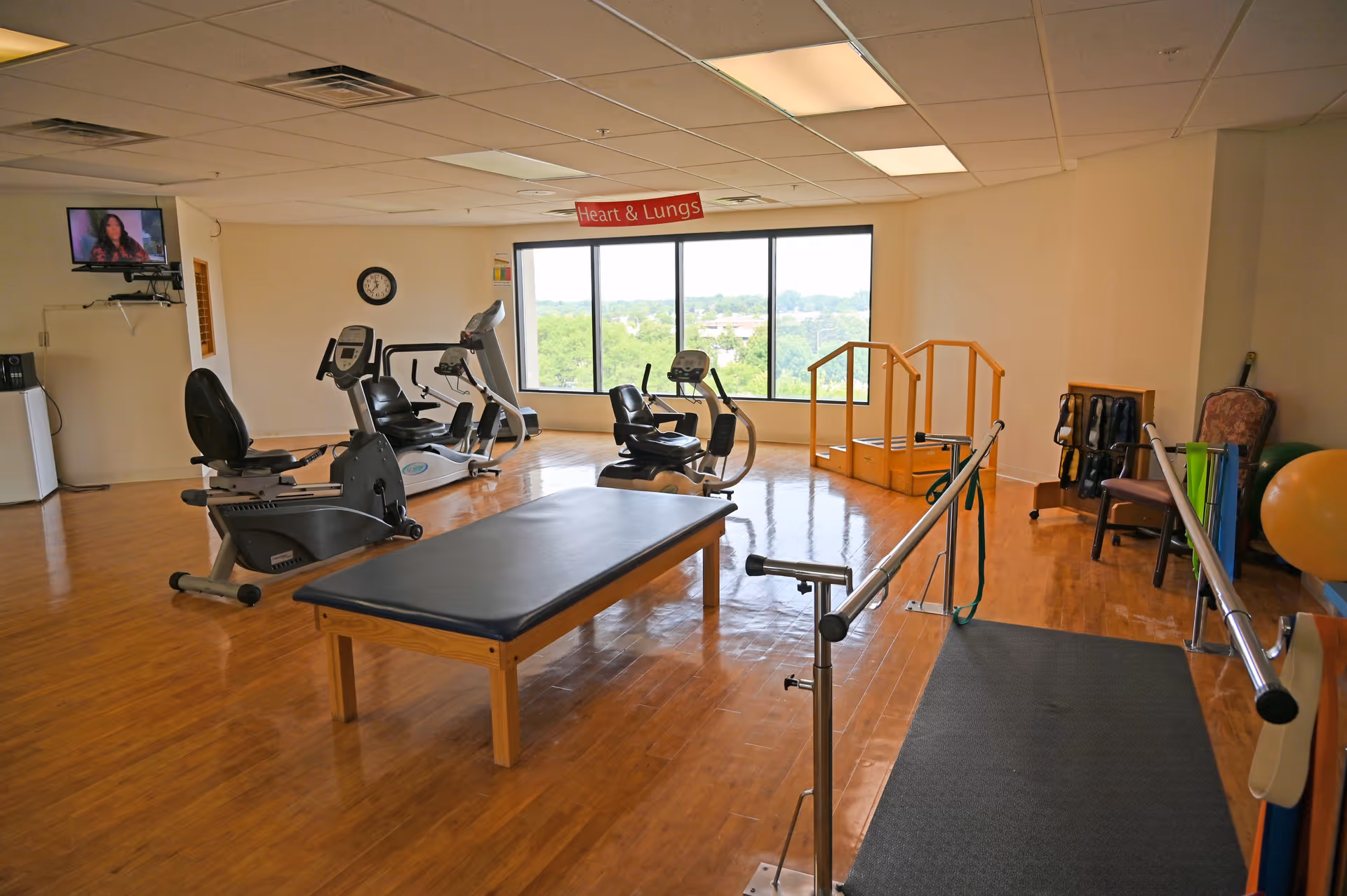 A spacious rehabilitation or physical therapy room with exercise equipment including stationary bikes, parallel bars for walking practice, a padded therapy table, and a set of stairs for mobility training. The room has large windows with a view of greenery outside, a wall-mounted TV, and a sign hanging from the ceiling that reads 'Heart & Lungs'.