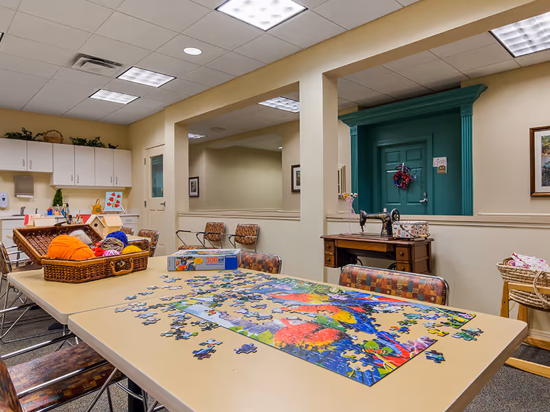 A well-lit activity room with a large table covered in a colorful jigsaw puzzle in progress. The table also holds a basket filled with yarn balls and craft supplies. The room has beige walls, patterned chairs, white cabinets, and a vintage sewing machine on a wooden table against the wall. A green door with a wreath is visible in the background.
