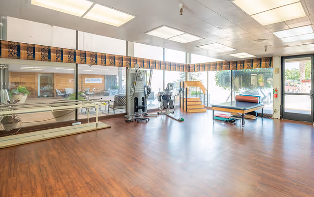 A spacious rehabilitation or physical therapy room with wooden flooring, large windows allowing natural light, exercise equipment including a multi-gym machine, stationary bike, therapy table with a bolster, and parallel bars for walking exercises. There is a door leading outside and some seating visible through the glass partition.