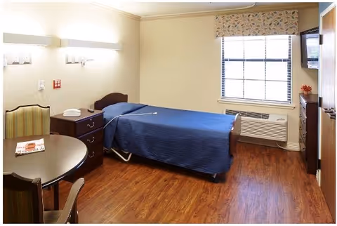 A simple, clean bedroom in a senior living facility with a single bed covered in a blue blanket, a wooden nightstand with a telephone, a round table with two chairs, a window with patterned curtains, a wall-mounted TV, and wood flooring.