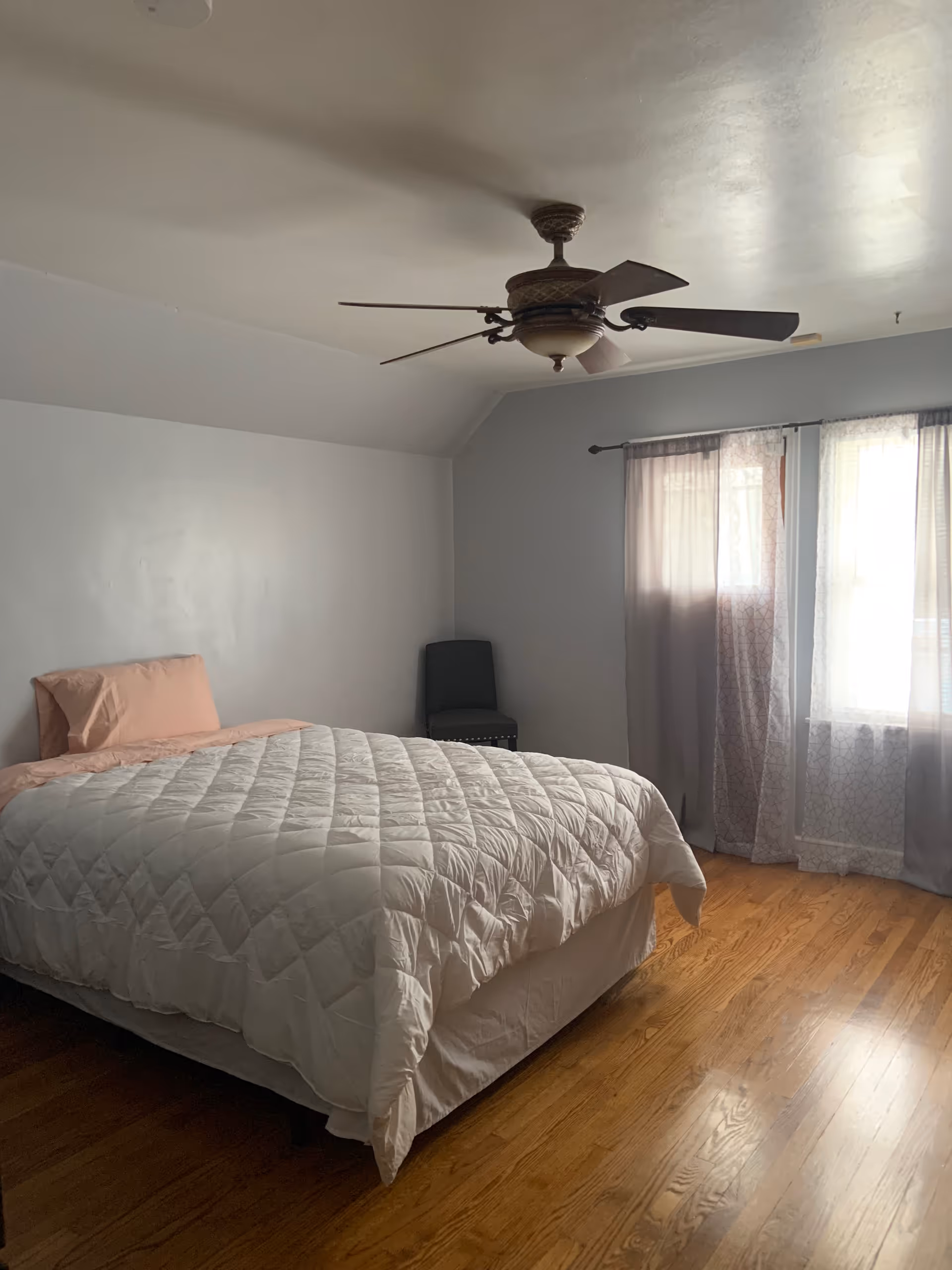 A simple bedroom with a bed covered in a white quilt and a pink pillow, a dark chair in the corner, wooden floor, a ceiling fan, and two windows with sheer curtains letting in natural light.