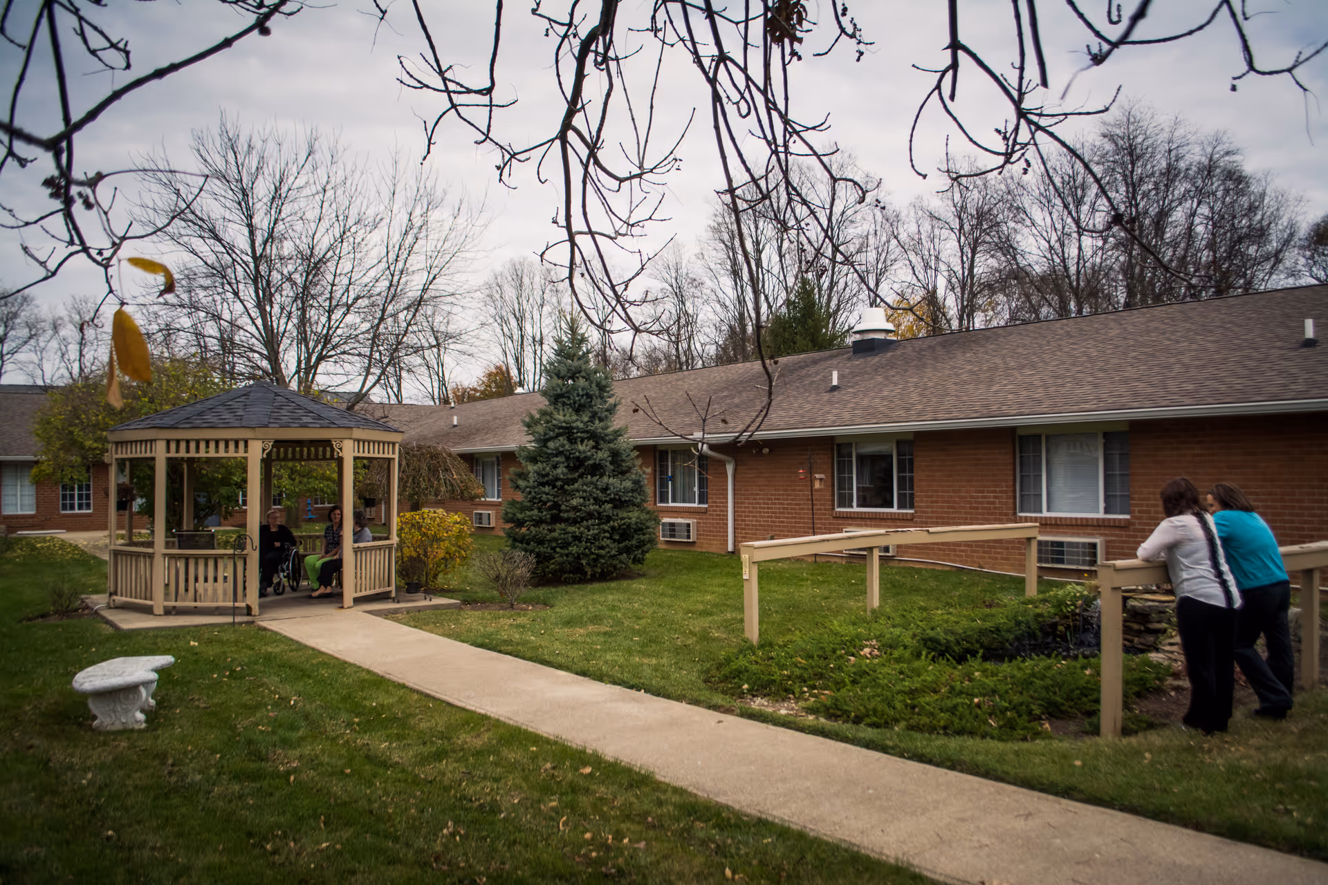 Outdoor area of a senior living facility with a wooden gazebo where two people are seated, one in a wheelchair. Two other people are standing and leaning on a wooden railing near a garden bed. The building is a single-story brick structure with several windows and air conditioning units. Leafless trees and a cloudy sky are visible in the background.