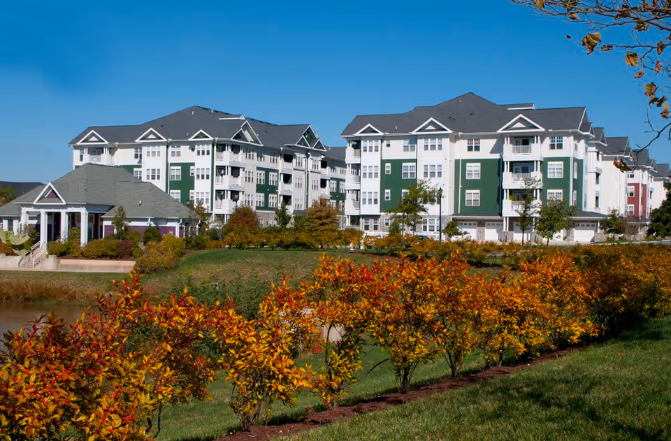 Exterior view of The Willows at Victoria Falls senior living facility showing multiple multi-story buildings with white and green facades, surrounded by landscaped grounds with bushes and trees under a clear blue sky.