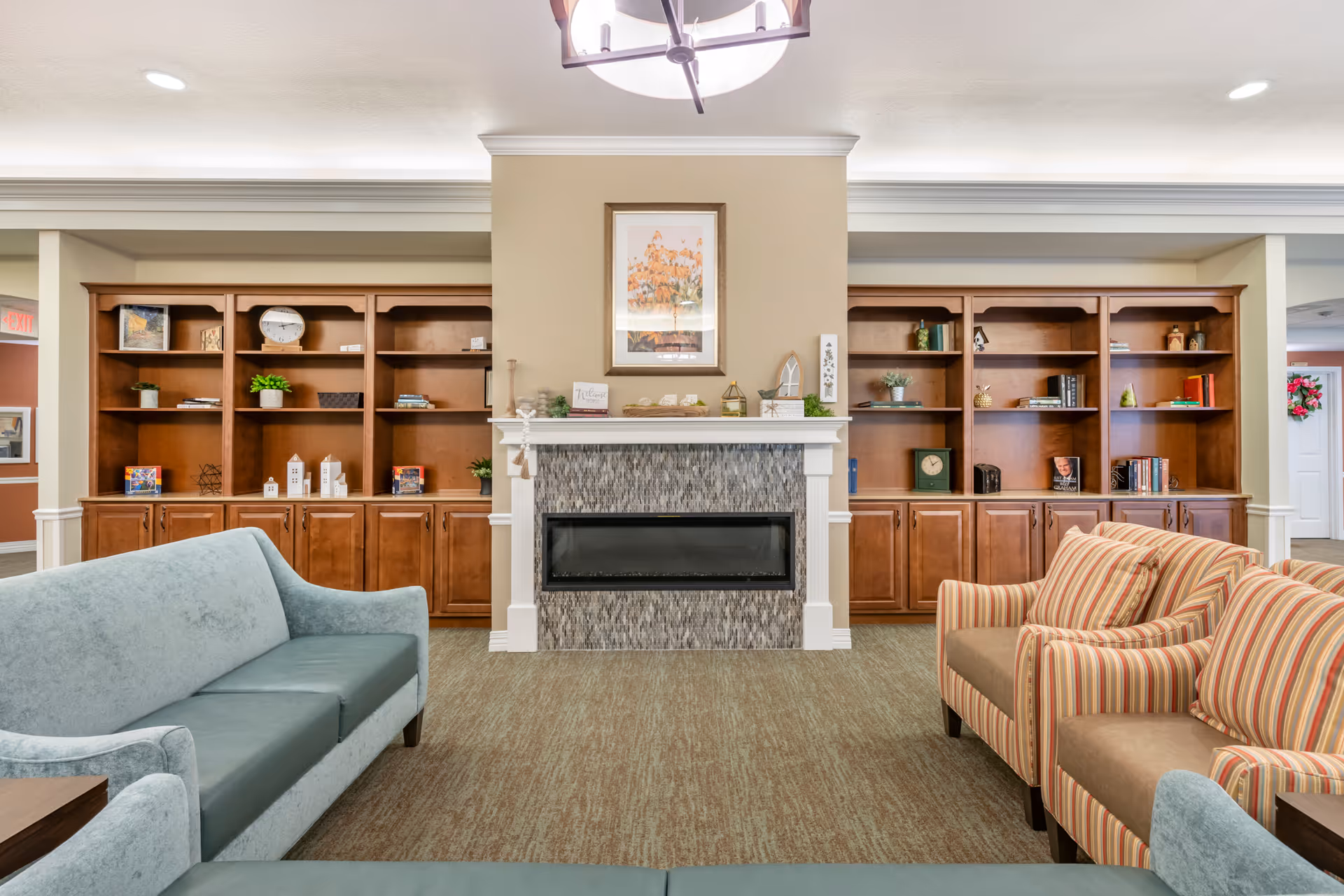 A cozy living room area in a senior living facility featuring a modern electric fireplace with a tiled surround and white mantel. On either side of the fireplace are wooden built-in shelves and cabinets decorated with books, plants, clocks, and small decorative items. The room has a light blue sofa and two striped armchairs arranged facing each other, with a soft carpeted floor and neutral wall colors. A framed picture hangs above the fireplace, and a ceiling light fixture is visible.