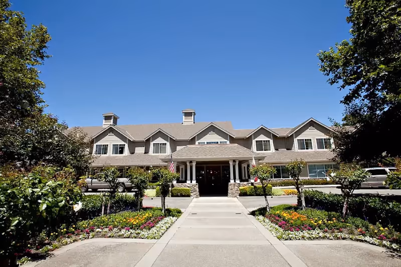 Front exterior view of a two-story senior living facility building with a peaked roof, multiple windows, and a covered entrance supported by columns. The building is surrounded by well-maintained landscaping including trees, bushes, and colorful flower beds under a clear blue sky.