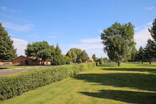 A well-maintained outdoor area with green grass, trimmed hedges, and several trees under a clear blue sky. In the background, there are single-story brick buildings along a paved road.