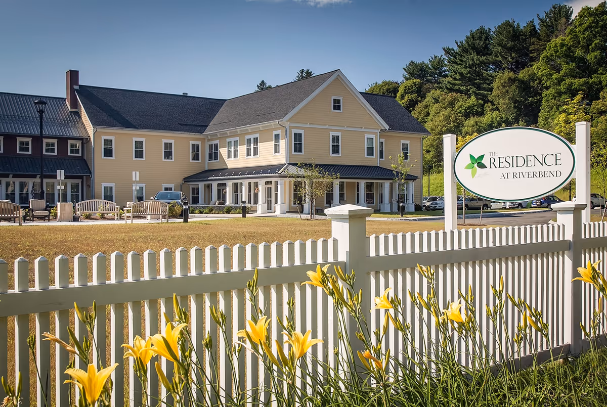 Exterior view of The Residence at Riverbend, a large two-story yellow building with white trim, surrounded by a white picket fence and yellow flowers in the foreground, with trees and a clear blue sky in the background.