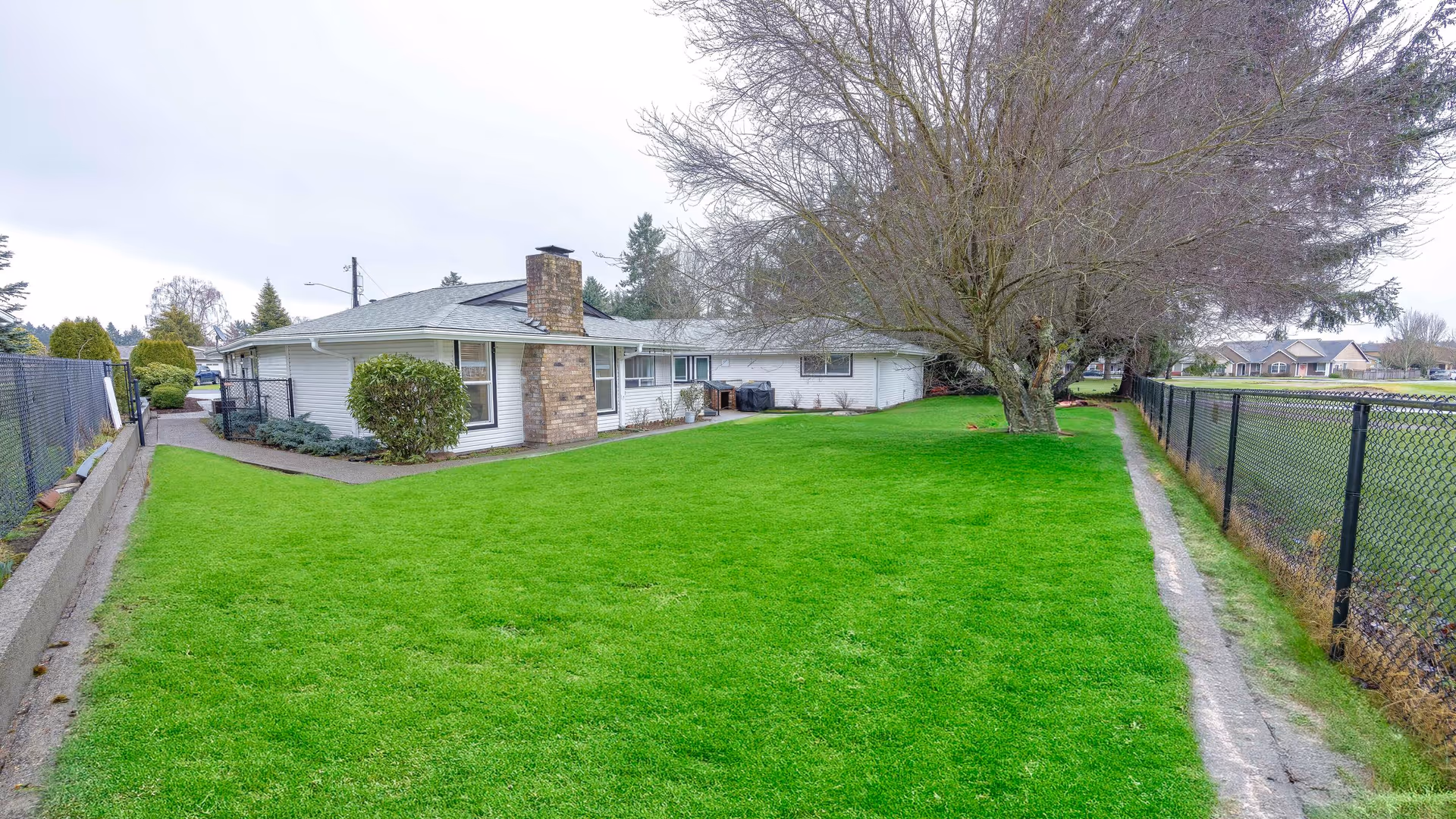 Single-story ranch-style building with a large green lawn, a leafless tree near the house, and a chain-link fence on the right.